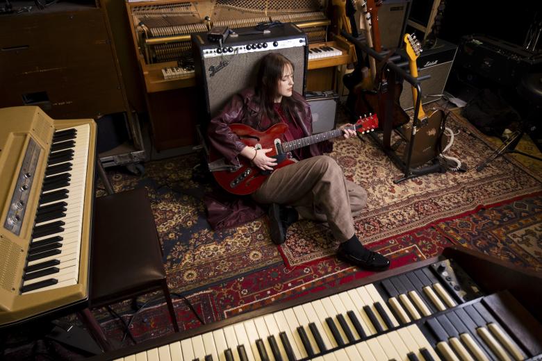 Pia Geffroy zit op de vloer terwijl ze tegen een versterker leunt en een rode gitaar vasthoudt.